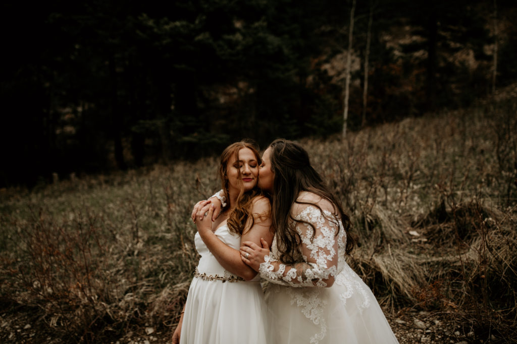 woman kissing new wife on cheek in a field wearing their wedding dresses