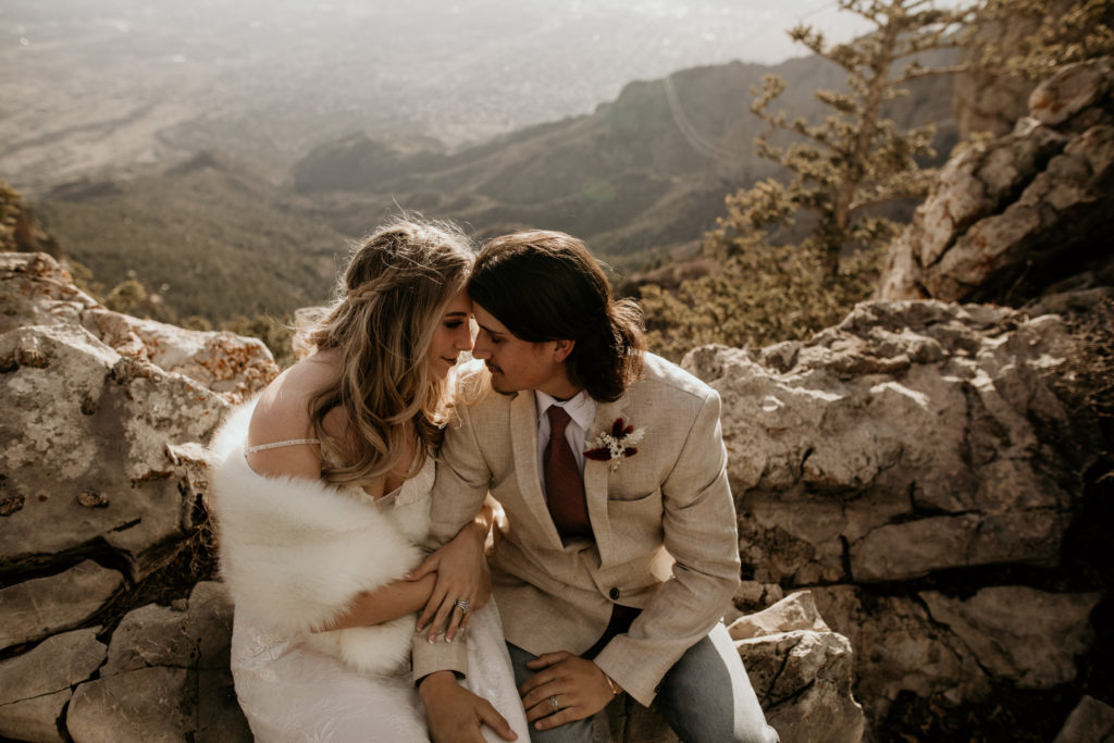 husband and wife sittins on a rock on top of the Sandia mountains overlooking Albuquerque