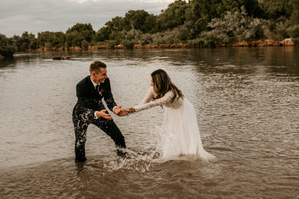 husband and wife splashing water in a river in their wedding clothes