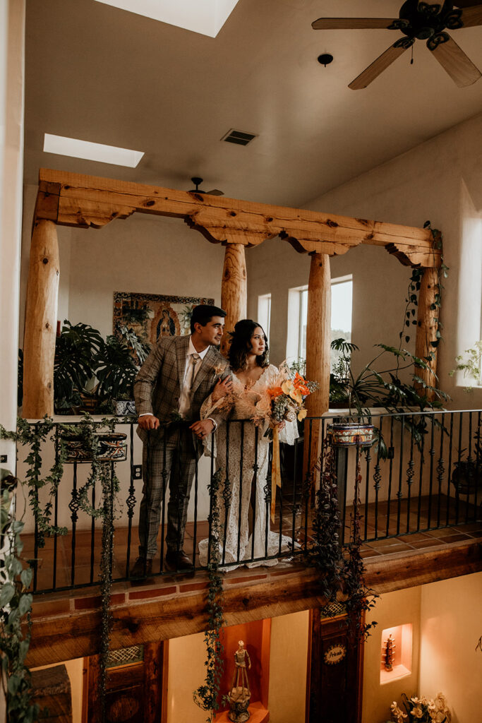 Newlyweds standing on an indoor balcony surrounded by vining plants. 