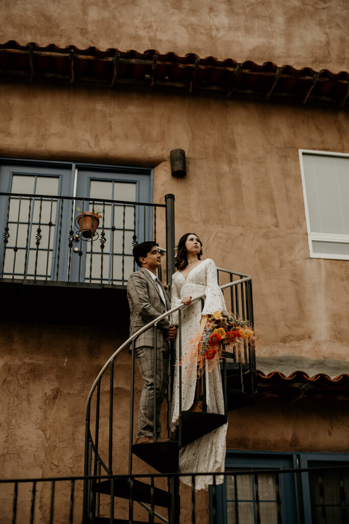 Newlyweds standing on a spiral staircase in their wedding attire outside of an adobe style building with turquoise doors. 