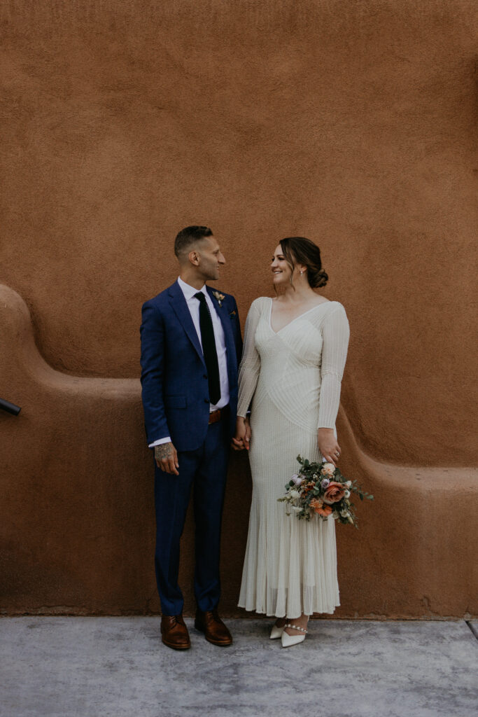 Bride and groom standing together outside of an adobe style building holding hands. 