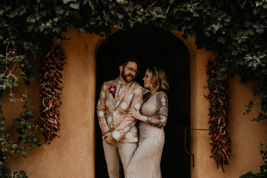 Bride and groom standing together in a doorway surrounded by red chile ristras at a Santa Fe wedding venue. 